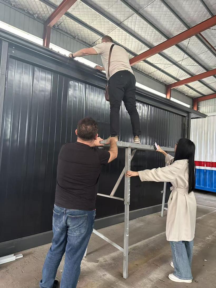 Client climbing ladder to inspect roof edge connections on dark-clad container building module inside factory